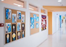 A long empty school corridor with children’s drawings on the wall. Classroom doors with classes going on inside.