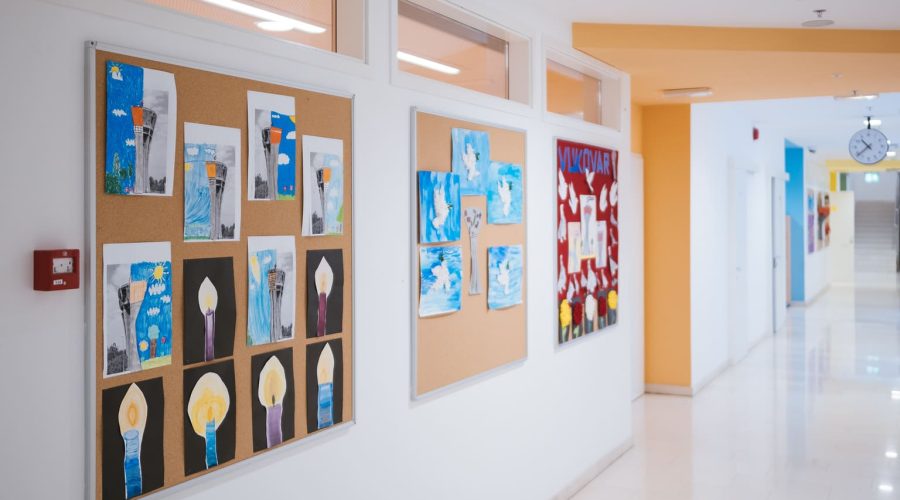 A long empty school corridor with children’s drawings on the wall. Classroom doors with classes going on inside.