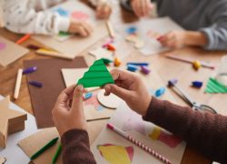 A primary school student cutting green paper at school, while other students are sitting at the desk cutting other coloured papers to create new images and shapes.