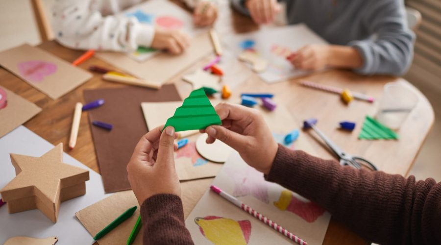 A primary school student cutting green paper at school, while other students are sitting at the desk cutting other coloured papers to create new images and shapes.