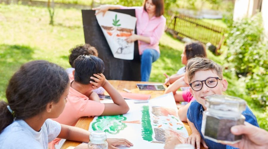 A group of secondary school pupils sitting at a table outside doing a school project regarding sustainable development, climate change and environmental awareness. The pupils are smiling and working together with their teacher sitting in the background explaining the lesson from a poster.