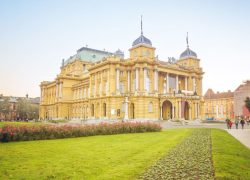 Croatian National Theatre in Zagreb, yellow neo-baroque building as seen from the front right side with green grass and flowers leading to the entrance on a bright sunny day. This photo is a link to Zagreb Croatia location page.