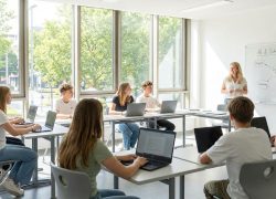 Students in a bright classroom working on laptops while a teacher leads a lesson on AI literacy and critical thinking.